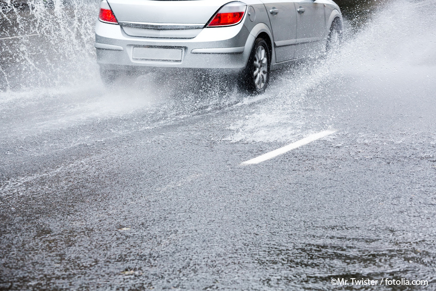 Mit wasser überflutete Straße, auf der ein fahrendes silbergraues Auto Spritzwasser erzeugt